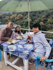 Trekking with Kids in the Annapurna Region: Family Adventure in Nepal 3 A man and two children are sitting at an outdoor table covered with a blue floral tablecloth, enjoying a meal under a green umbrella in a hilly area, resting after a morning of trekking with kids.
